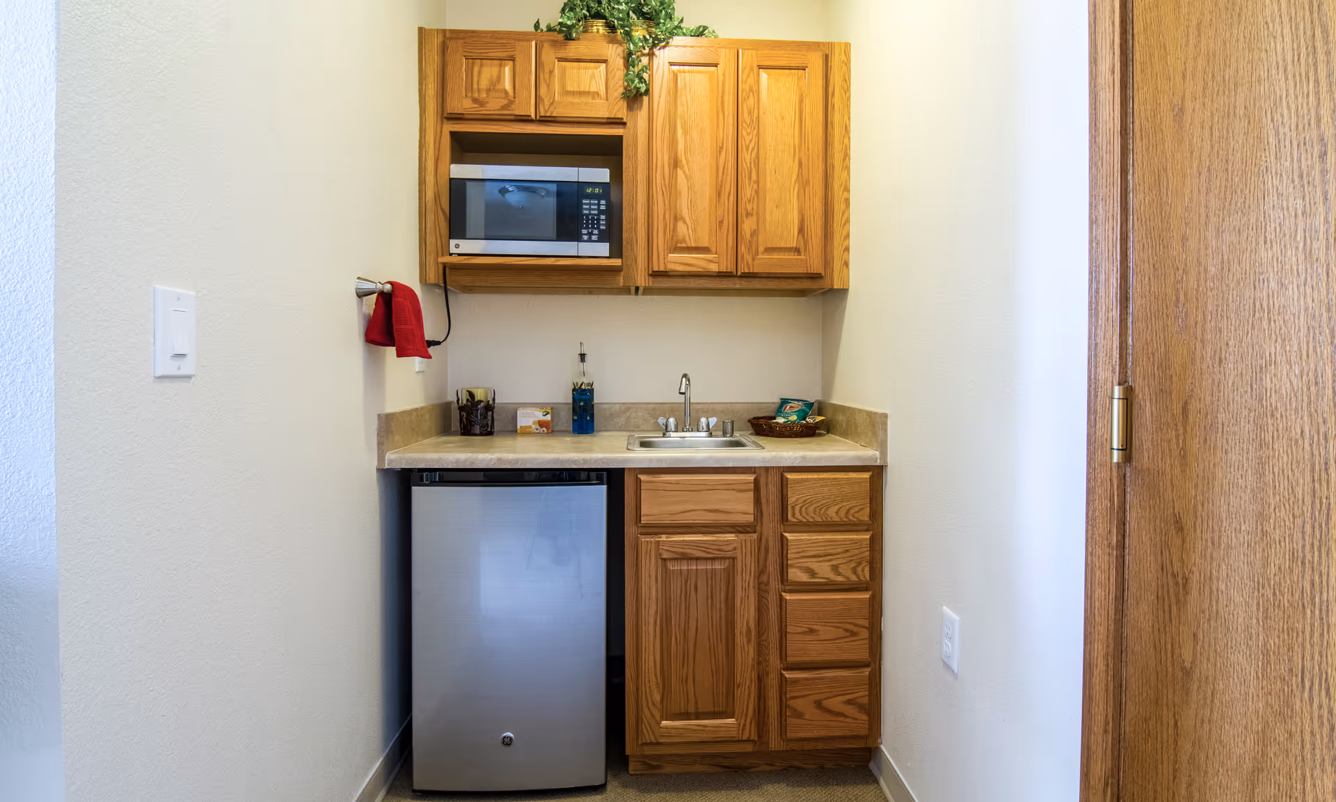 Small kitchenette area with wooden cabinets, a microwave, a mini refrigerator, a sink, and a countertop with a few items including a basket of snacks and a bottle of dish soap. A red towel hangs on a hook on the left wall.