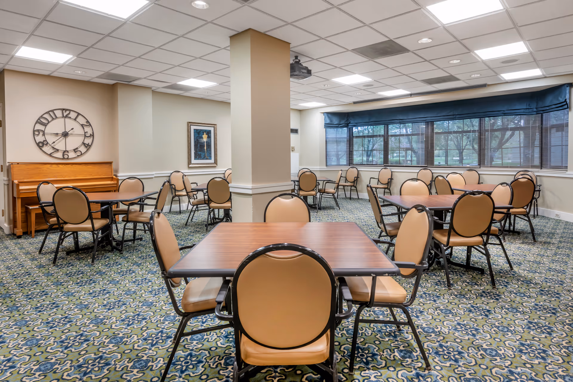 A spacious dining room with multiple square wooden tables surrounded by beige cushioned chairs with black frames. The room has a patterned carpet in shades of blue and green, a large window with blue valances, a wall clock, a framed picture, and a wooden piano against the wall.