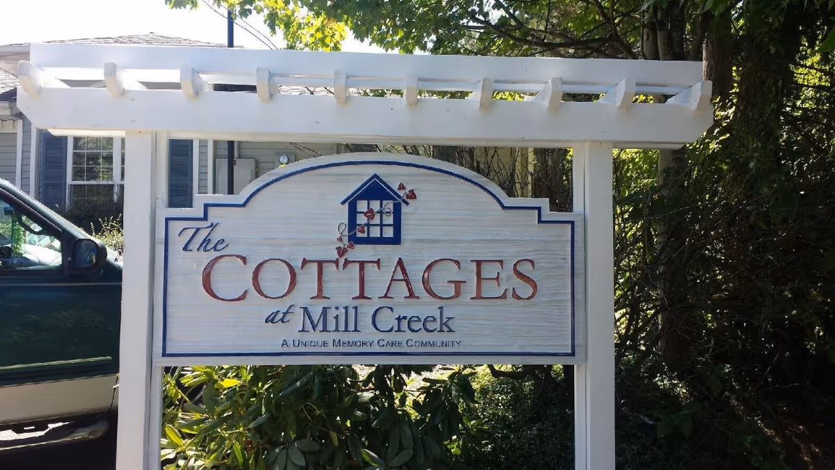 White wooden sign reading 'The Cottages at Mill Creek' mounted under a pergola in front of greenery and a house.