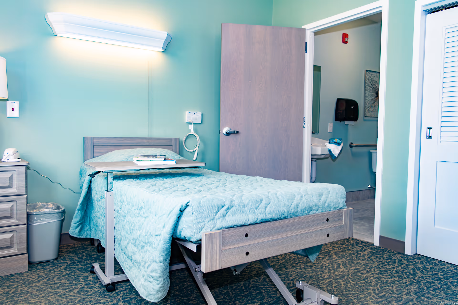 A senior living facility bedroom with a hospital-style adjustable bed covered with a light blue quilt. There is a bedside table with a lamp and a telephone, a trash can, and an overbed table with some items on it. The room has light green walls and carpeted floor. An open door reveals a bathroom with a sink, toilet, and handrails.