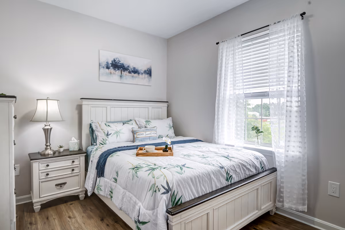 A bright bedroom with a white wooden bed frame and matching nightstand. The bed is made with white bedding featuring a green leaf pattern, and a wooden tray with a cup and small plant is placed on top. A lamp, tissue box, and small plant are on the nightstand. A window with sheer white curtains lets in natural light, and a painting of trees hangs above the bed.