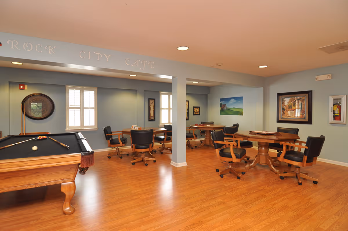 A spacious room with light blue walls and wooden flooring featuring a pool table on the left and several round wooden tables with black cushioned chairs on wheels. The room has windows with white shutters and framed artwork on the walls. Above the room entrance, the words 'Rock City Cafe' are displayed.