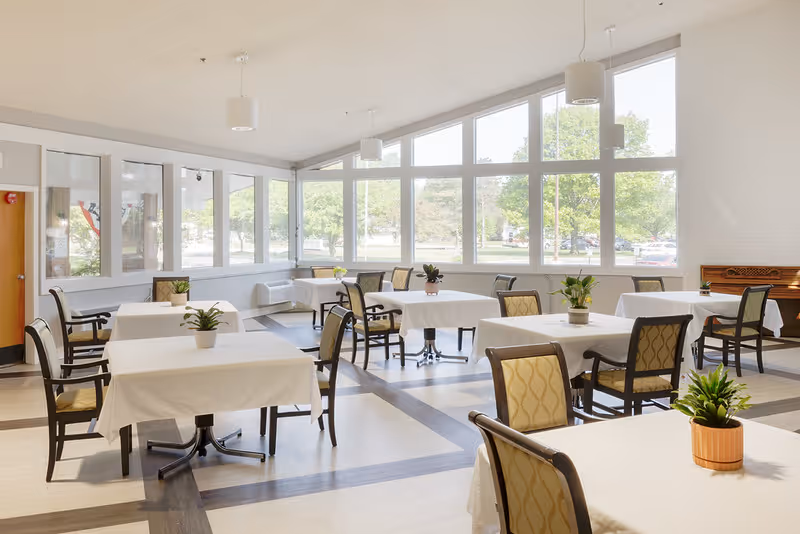 Bright dining room with tables covered in white tablecloths, potted plants on each table, and large windows letting in natural light.