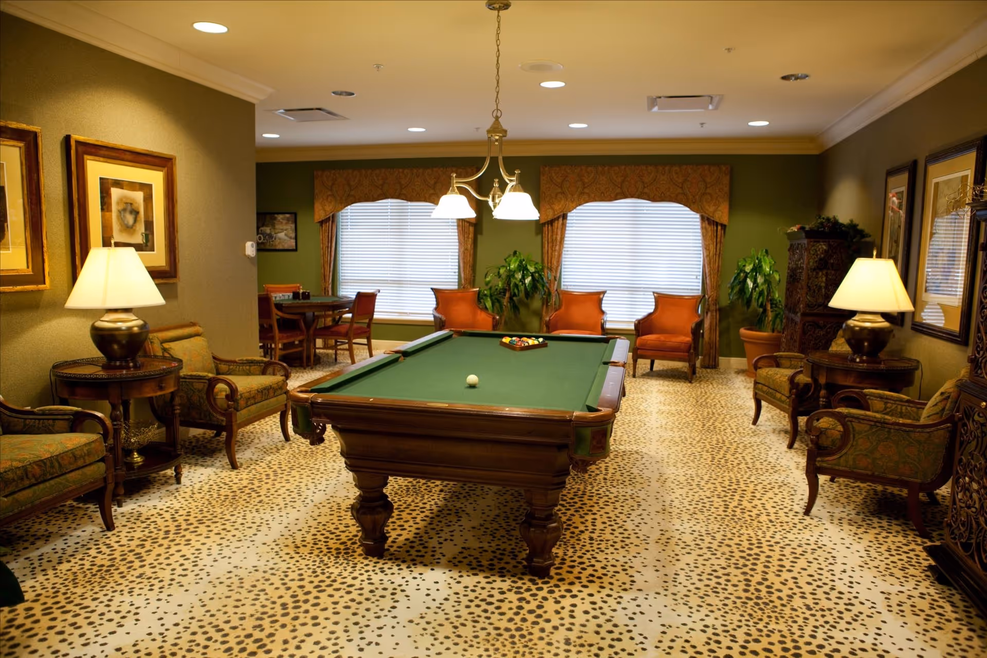 A cozy game room with a pool table in the center, surrounded by upholstered chairs and small tables with lamps. The room has green walls, patterned carpet, framed artwork, and two large windows with blinds and valances. A small dining table with chairs is visible in the background.