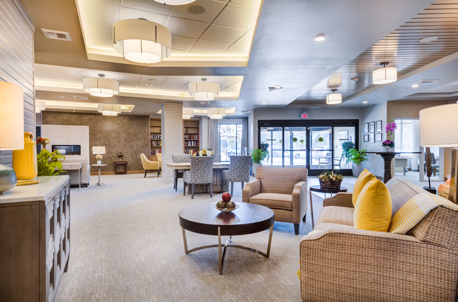 Spacious, well-lit communal lounge with sofas, armchairs, a round coffee table, dining tables, and glass entrance doors.