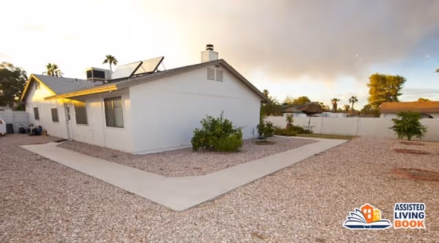 Exterior view of a single-story white building with solar panels on the roof, surrounded by a gravel yard with a concrete walkway and some small plants and trees under a partly cloudy sky.