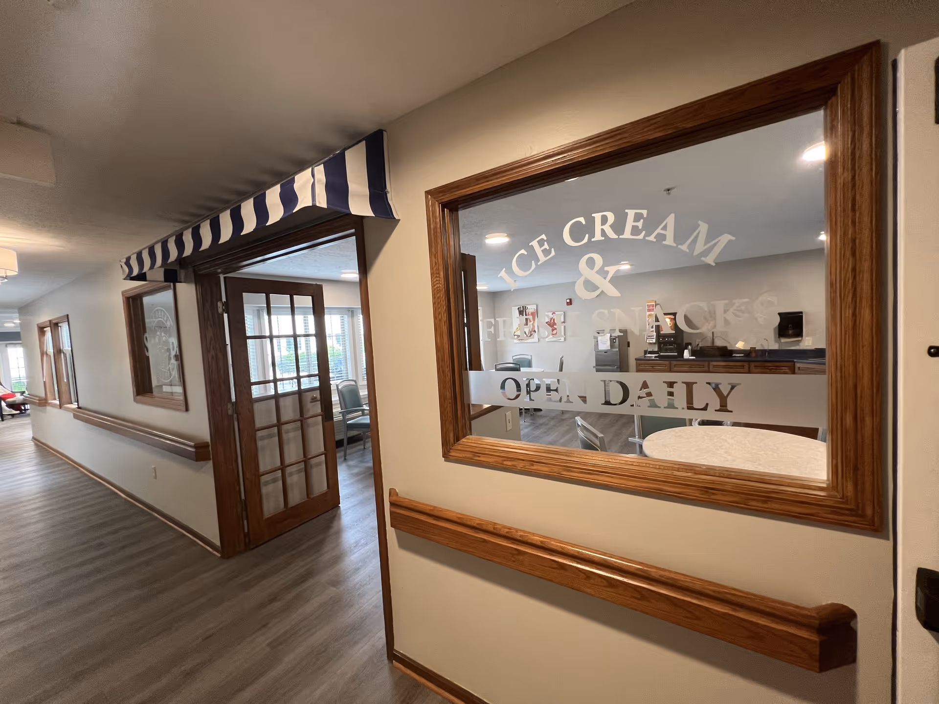 Interior hallway of a senior living facility with wood-trimmed windows and doors. One window has frosted text that reads 'ICE CREAM & FRESH SNACKS OPEN DAILY'. Inside the room visible through the window are chairs and tables, with a striped blue and white awning above the door.