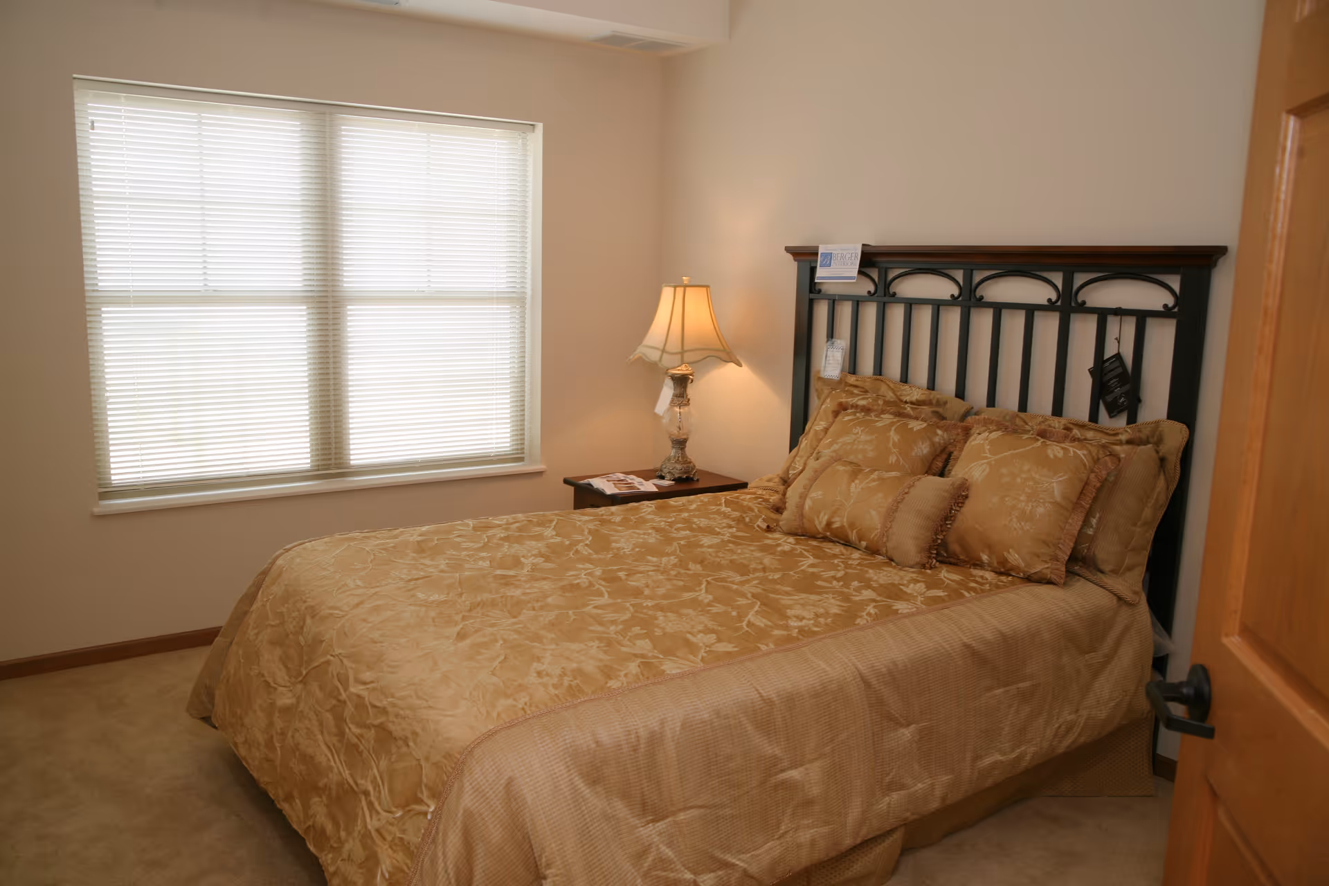 A bedroom with a neatly made bed covered in a gold patterned bedspread and matching pillows. There is a wooden headboard with decorative metalwork, a nightstand with a lamp, and a window with closed blinds letting in natural light.