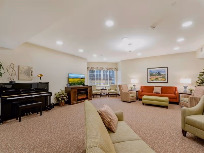 A spacious and well-lit living room with beige walls and carpeted floor. The room features multiple seating options including a green sofa with a striped pillow, a red sofa with a pillow, several armchairs, and a green ottoman. There is a black upright piano on the left side with a small bench, a wooden TV stand with a flat-screen TV displaying a field of sunflowers, and a large window with floral valance letting in natural light. The room is decorated with framed artwork, table lamps, plants, and a decorative musical note sculpture.