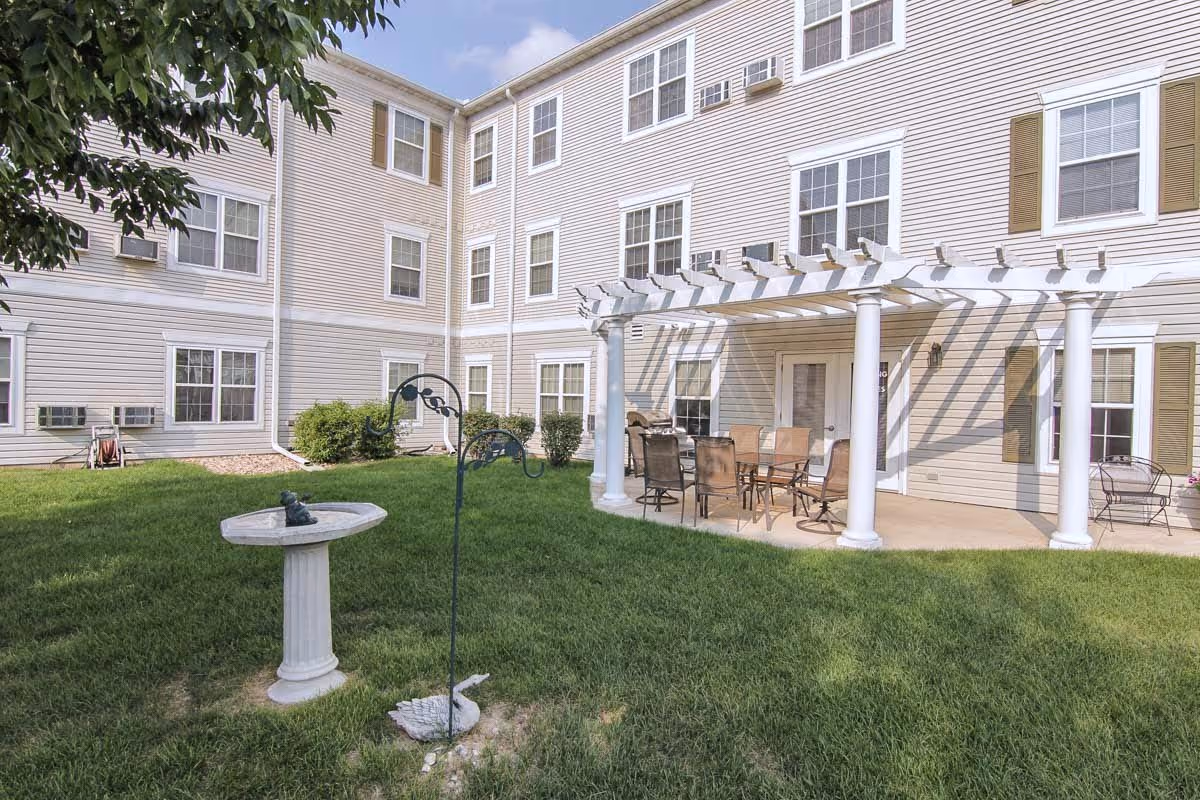 Outdoor courtyard area of Fox Meadow Senior Apartments featuring a green lawn, a birdbath, a decorative bird feeder, and a patio with a white pergola and outdoor dining furniture. The building exterior is beige with multiple windows and brown shutters.