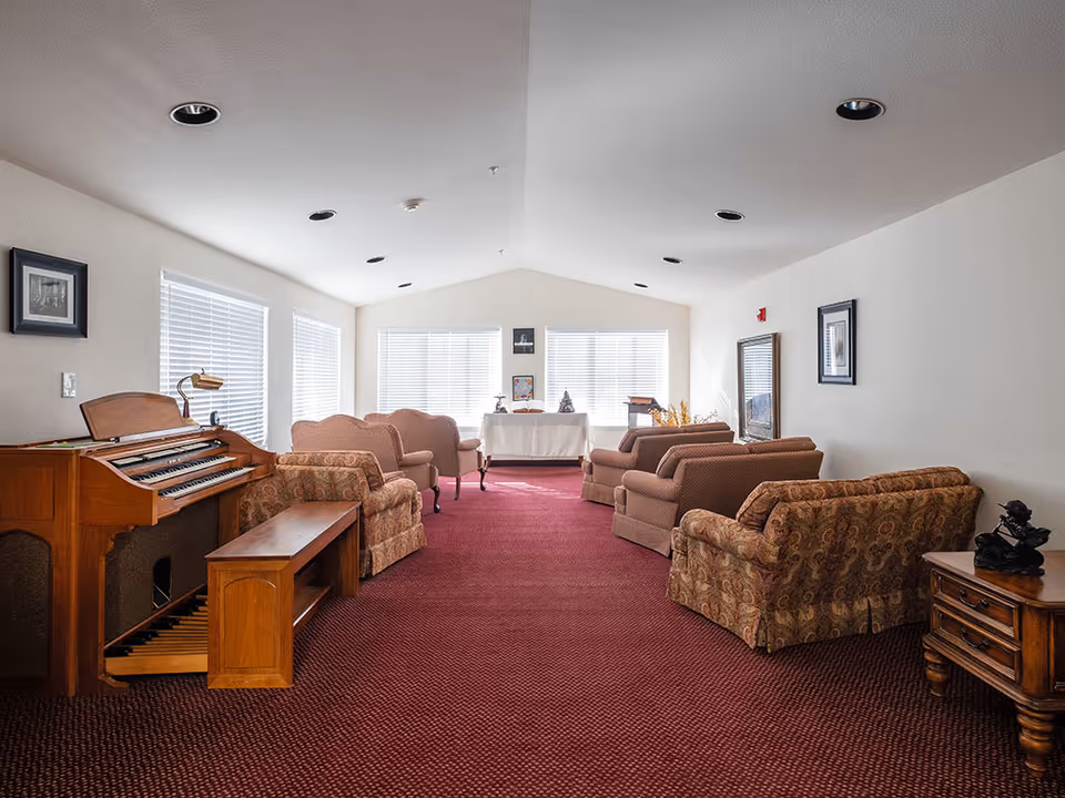 A spacious living room with a red carpet, multiple upholstered armchairs and sofas arranged in a conversational layout, a wooden organ with a bench on the left side, and large windows with white blinds letting in natural light. The walls are decorated with framed pictures and a mirror, and there is a small wooden side table with a decorative sculpture on the right.