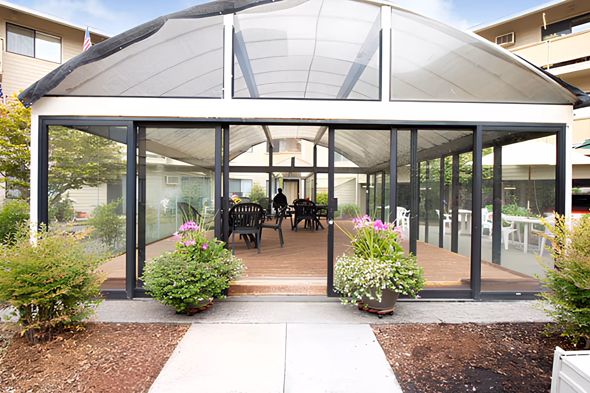 A glass-enclosed patio area with a curved roof, featuring several black chairs and tables inside. There are two large flower pots with pink and white flowers placed symmetrically on either side of the entrance. Surrounding the patio are bushes and a walkway leading up to the entrance. The background shows parts of the building exterior and some greenery.