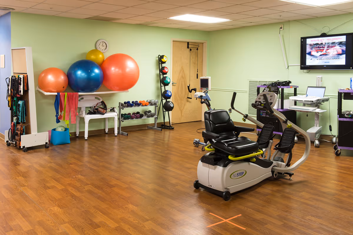 A physical therapy or exercise room with wooden flooring and light green walls. The room contains various exercise equipment including a recumbent exercise bike, colorful exercise balls on a shelf, resistance bands hanging on a rack, dumbbells on a rack, and a wall-mounted TV screen displaying information. There is also a clock on the wall and a wooden door in the background.
