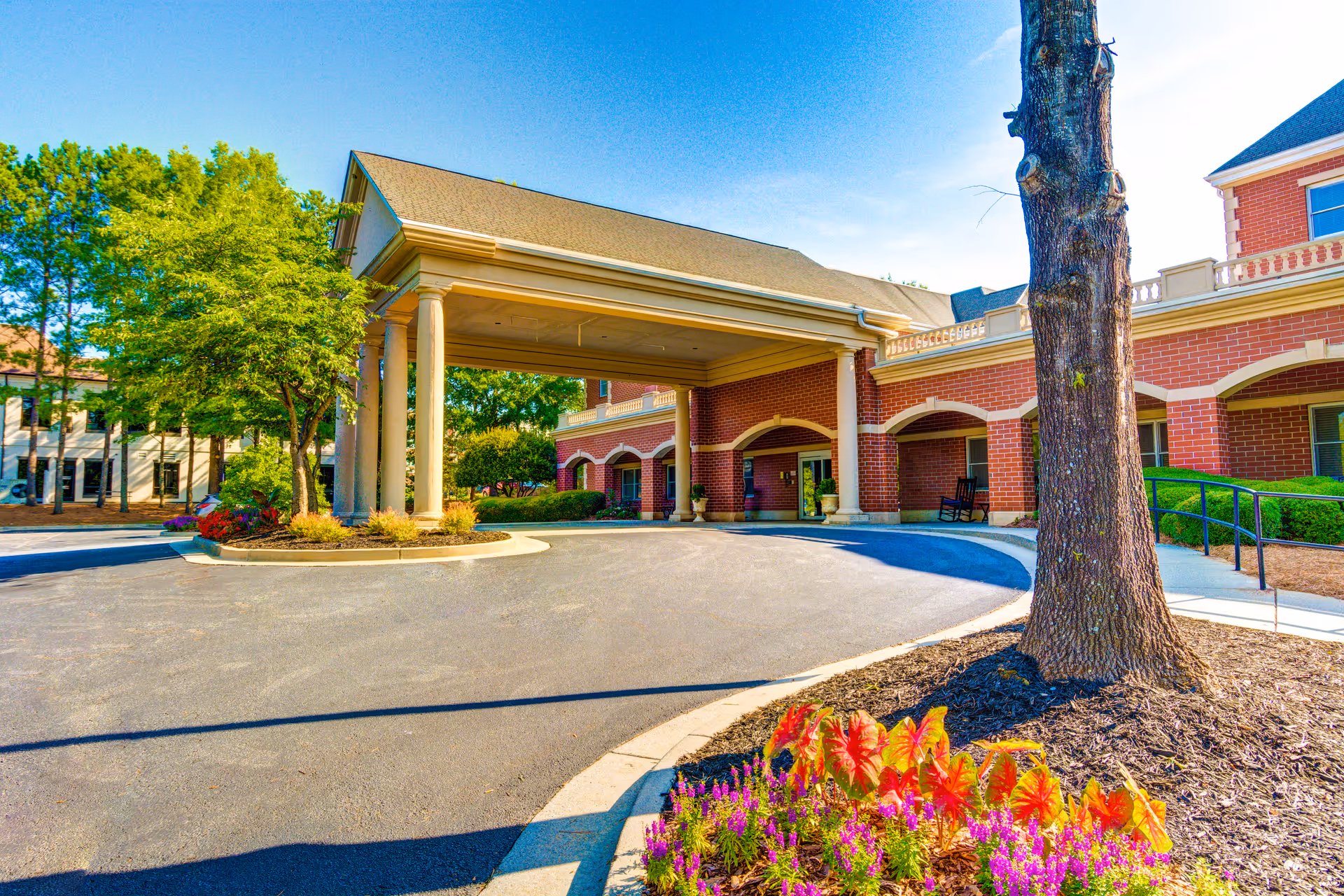 Entrance area of Dunwoody Place facility featuring a covered driveway with large columns, red brick building facade, landscaped flower beds with colorful flowers, trees, and clear blue sky.
