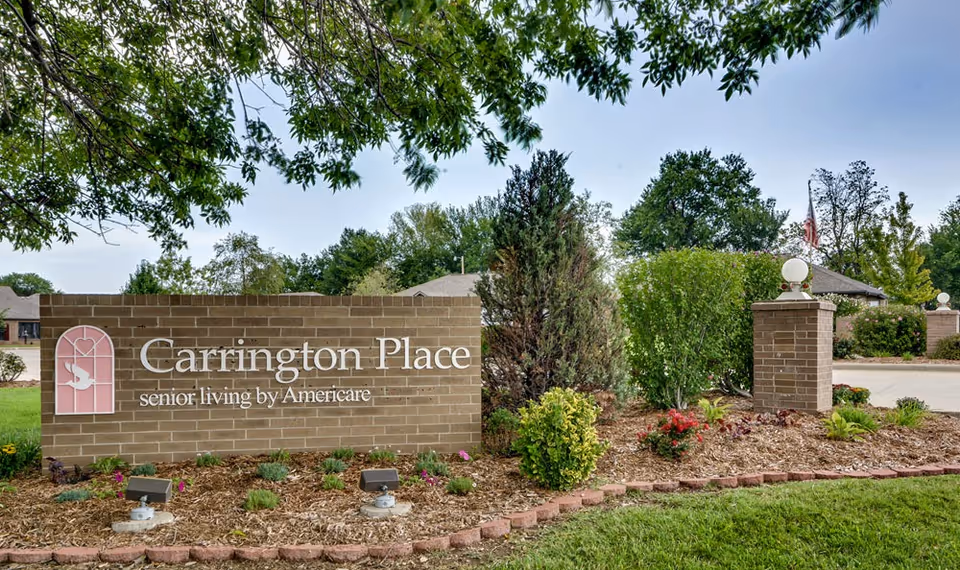 Outdoor view of the entrance sign for Carrington Place senior living by Americare, featuring a brick wall with the facility name and logo, surrounded by landscaped bushes, flowers, and trees under a clear sky.