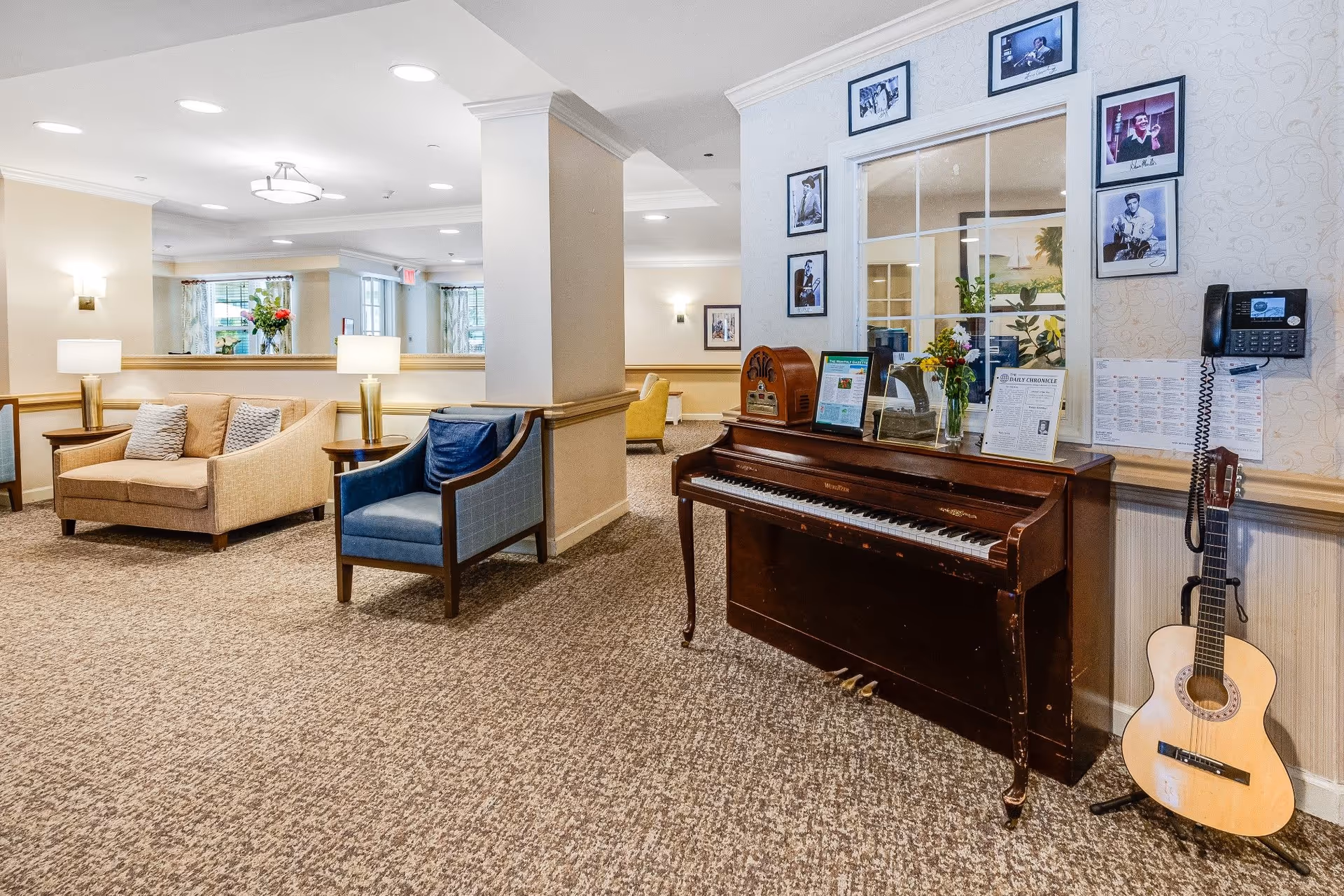 A cozy senior living facility common area with beige carpet and light-colored walls. The room features a wooden upright piano with framed photos above it, a vintage radio, a vase with flowers, and a guitar on a stand next to a wall-mounted telephone. Comfortable seating includes a beige loveseat with pillows and a blue armchair. The space is well-lit with ceiling lights and table lamps, and windows with curtains are visible in the background.