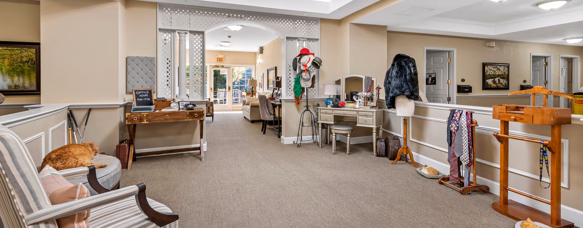 Interior hallway of a senior living facility with beige walls and carpeted floor. On the left, a striped armchair with a sleeping orange cat on a round ottoman. A wooden desk with a small chalkboard displaying office hours is against the wall. In the center, a white lattice archway leads to a sitting area with sofas and a glass door to the outside. On the right, a vanity table with a mirror and various accessories, a coat stand with hats, a mannequin with a black fur coat, and a wooden valet stand with ties hanging. Another orange cat is curled up on a small bed on the floor.