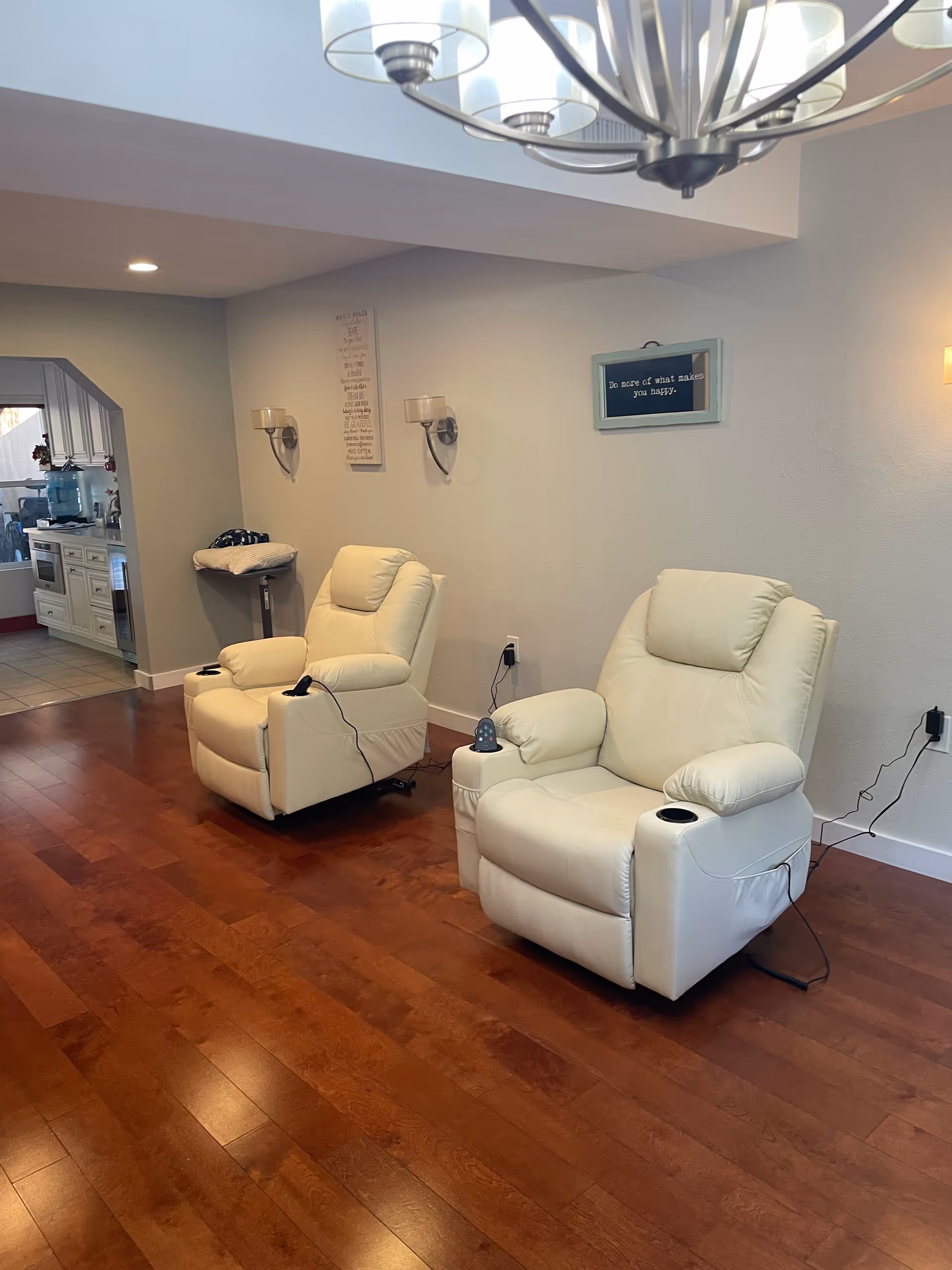 Two white recliner chairs with cup holders and remote controls on a wooden floor in a room with light-colored walls. A kitchen area is visible in the background through an open doorway. Wall decorations include a framed quote and two wall sconces.