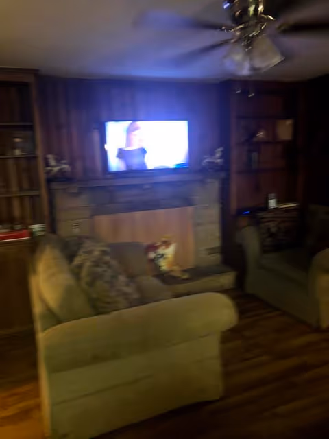 A cozy living room with two beige armchairs facing a wall-mounted television above a fireplace. The room has wooden paneling on the walls, built-in shelves on either side of the fireplace, and a ceiling fan with lights. The floor is wooden, and there are some decorative items on the shelves and mantel.