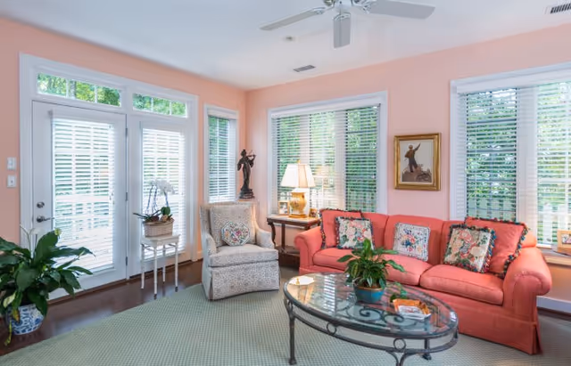 Bright living room with a coral sofa, patterned armchair, glass coffee table, and several windows with blinds.