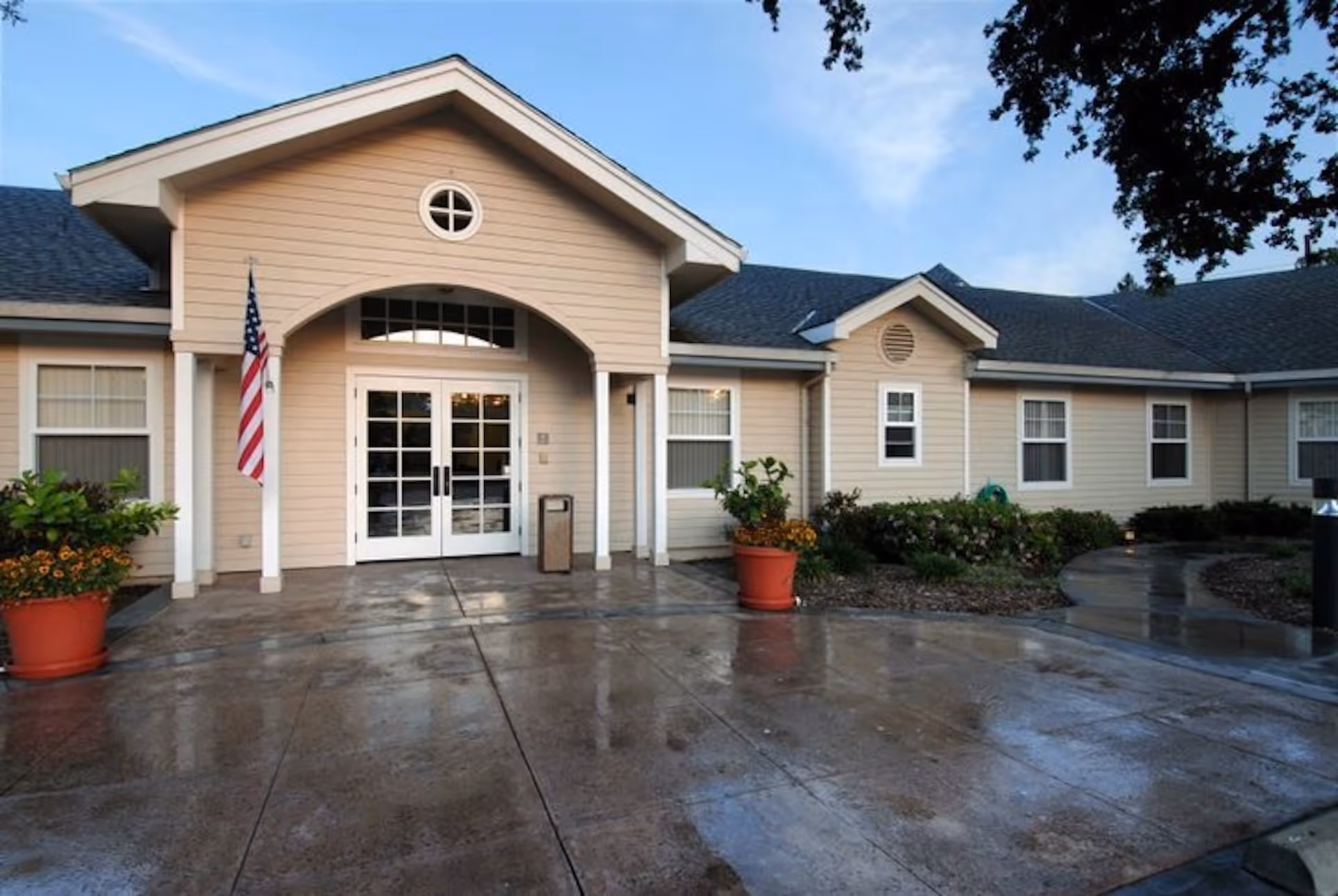 Exterior front entrance of a single-story building with beige siding, white trim, and a dark shingled roof. The entrance features double glass doors under an arched porch with a small round window above. There are two large potted plants with flowers on either side of the entrance, an American flag mounted on a white column, and a wet concrete walkway leading to the door. The sky is clear with some clouds and there are trees partially visible on the right side.