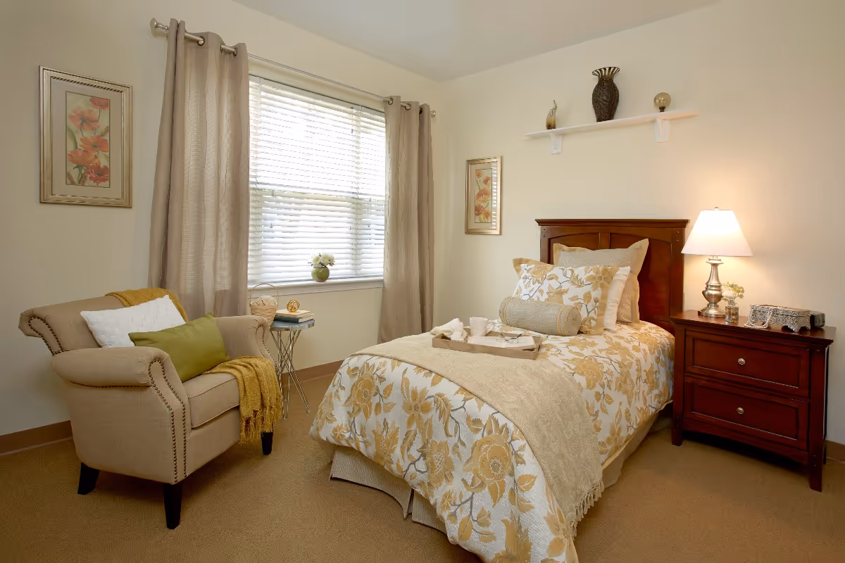 A cozy bedroom with a single bed covered in a floral patterned bedspread in shades of beige and yellow. The bed has multiple pillows and a tray with cups on it. Next to the bed is a wooden nightstand with a lamp and decorative items. A beige armchair with cushions and a yellow throw blanket is positioned near a window with beige curtains. The walls are light-colored with framed floral artwork and a small shelf holding decorative pieces.