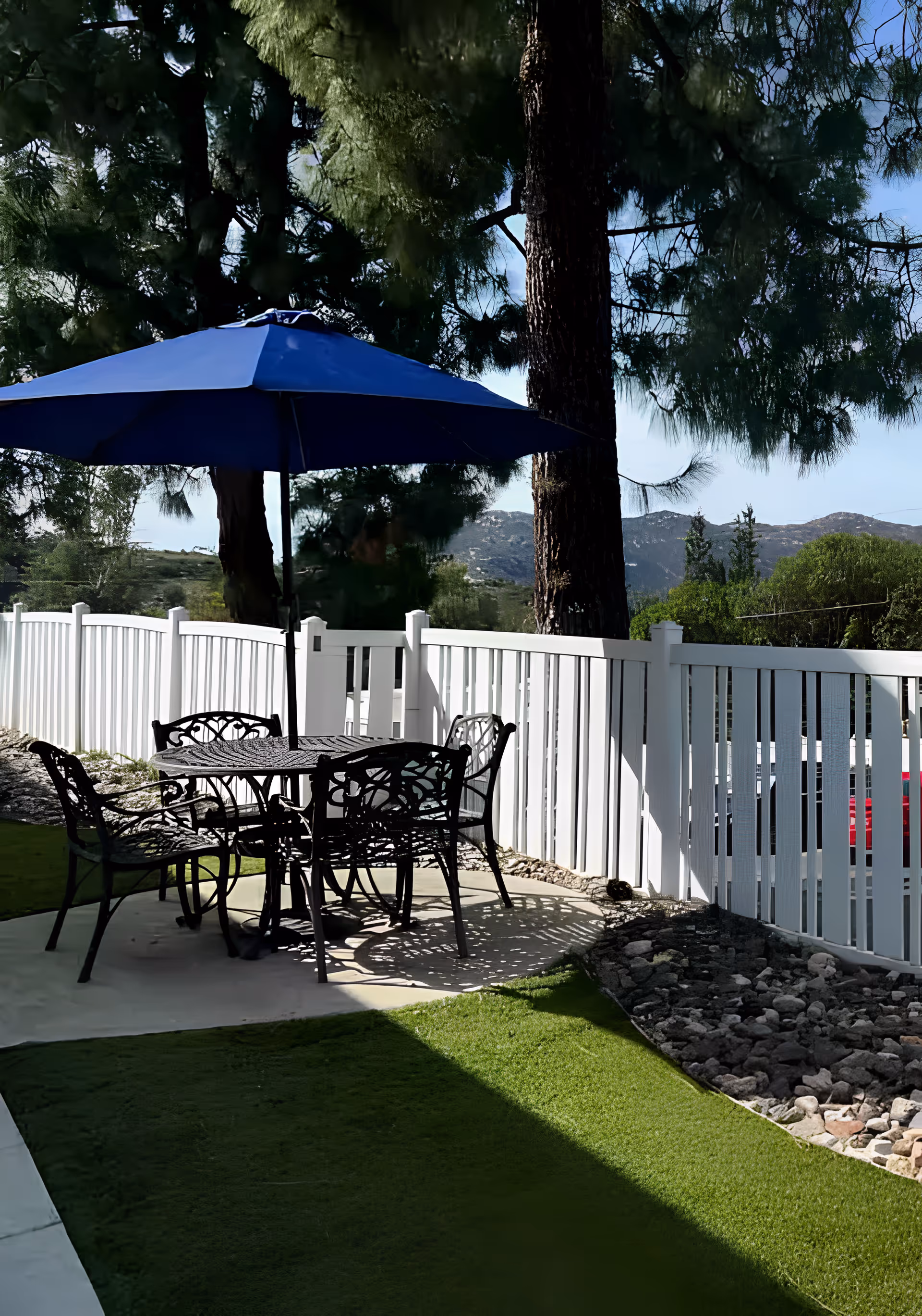 Outdoor patio area with a black metal table and four matching chairs under a large blue umbrella, surrounded by a white fence, with trees and mountains in the background.