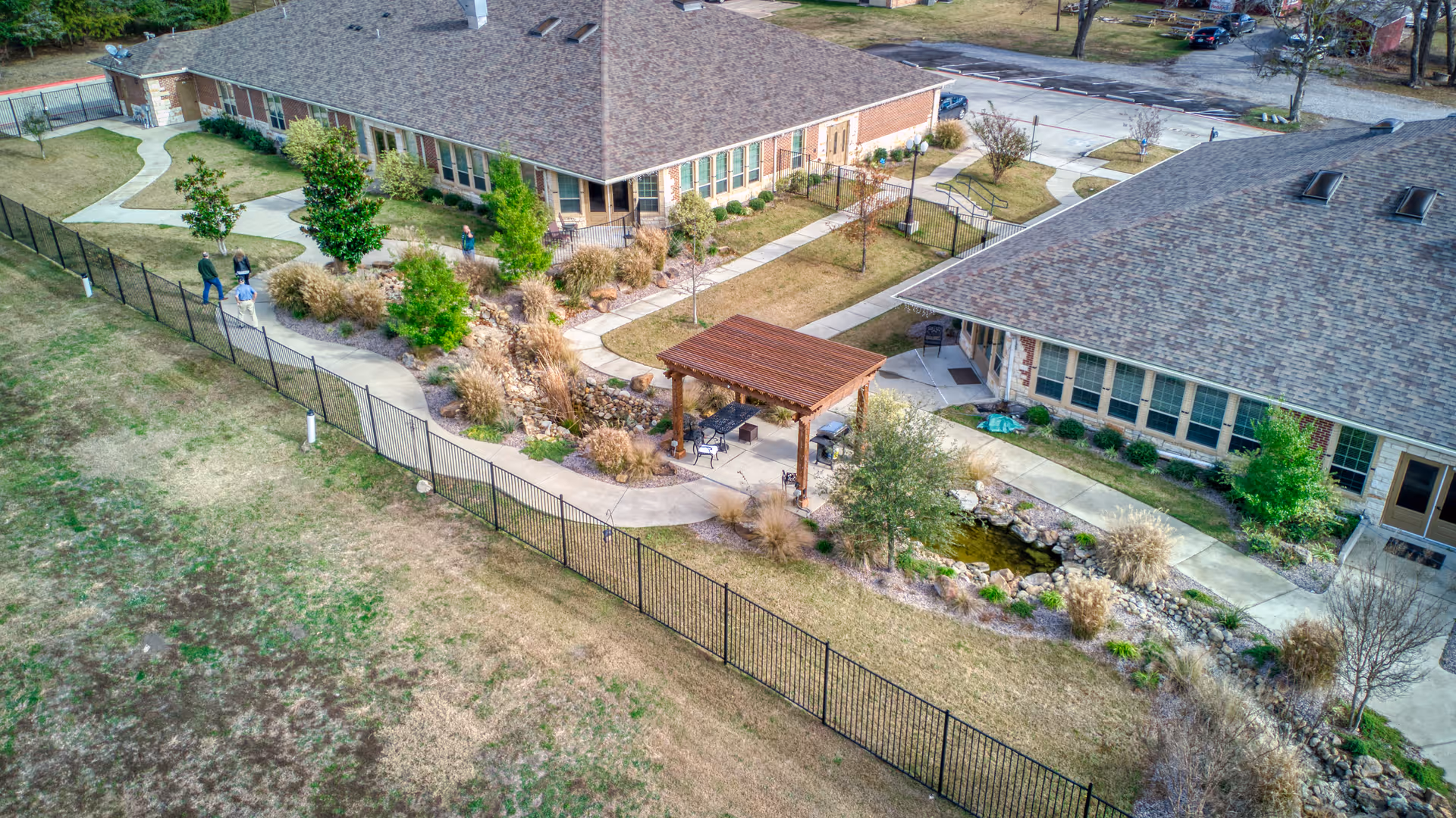 Aerial view of AvilaCare Assisted Living and Memory Care of Heath showing two single-story buildings with brown shingle roofs, surrounded by landscaped gardens and walking paths. There is a wooden pergola with seating near a small pond, and a black metal fence encloses the area. Several people are walking along the paths.