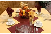 Dining table set for four with glass plates, white cups, folded burgundy and gold napkins, and a fall-themed centerpiece.