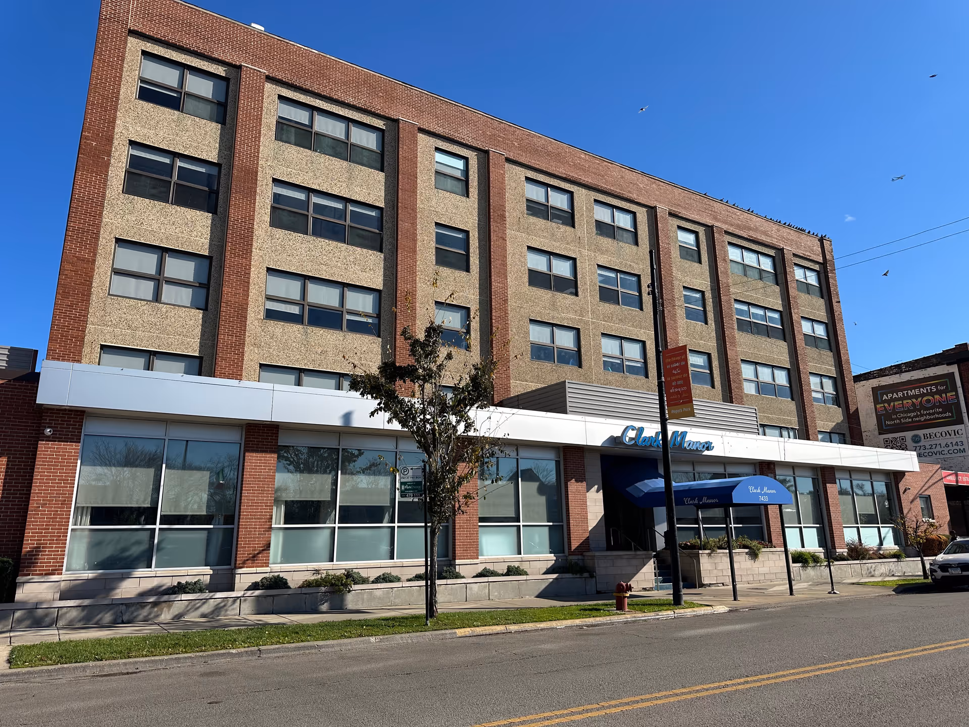 Exterior view of a multi-story brick and concrete building named Clark Manor with large windows and a blue awning over the entrance. The building is situated along a street with a sidewalk, a small tree, and a fire hydrant. The sky is clear and blue with a few birds flying.