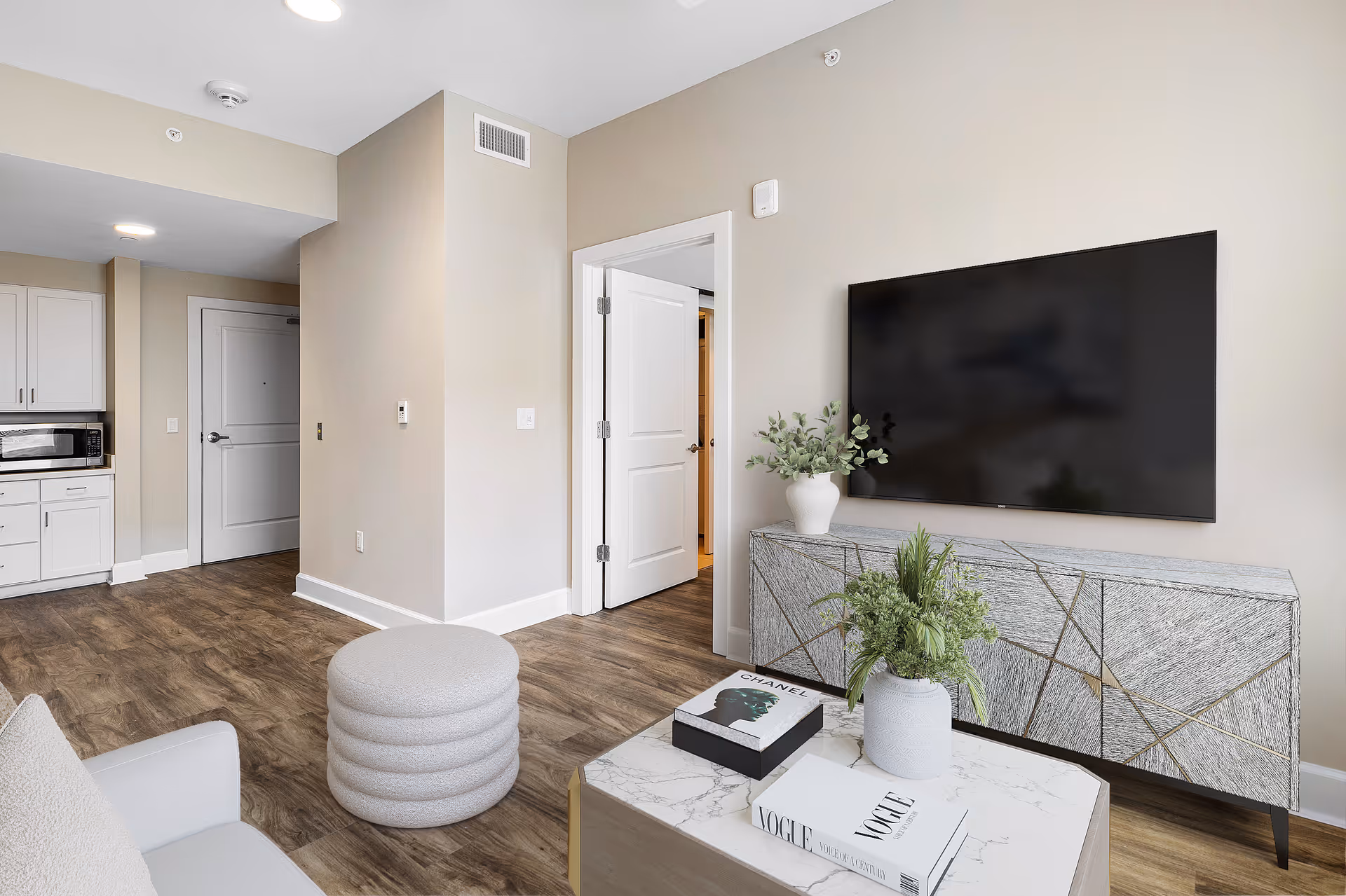 Modern living room area with a large flat-screen TV mounted on the wall above a stylish textured cabinet. A white vase with green plants sits on the cabinet. In front, there is a coffee table with two books and another white vase with green plants. To the left, part of a white sofa and a round, gray ottoman are visible. The room has wood flooring and beige walls, with a doorway leading to another room and a kitchen area with white cabinets and a microwave in the background.