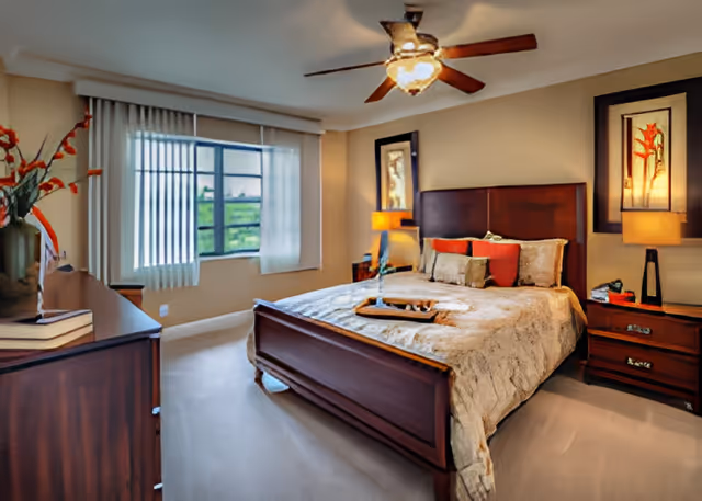 A well-lit bedroom with a large wooden bed featuring a beige comforter and multiple pillows. There are two wooden nightstands with lamps on either side of the bed, and framed artwork hanging above each nightstand. A ceiling fan with lights is mounted on the ceiling. A window with vertical blinds allows natural light into the room. A wooden dresser with books and a vase with flowers is positioned on the left side of the room.