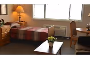 A small senior living bedroom with a single bed, nightstand and lamp, a window air conditioner, a table with chairs, and a coffee table with a potted plant.