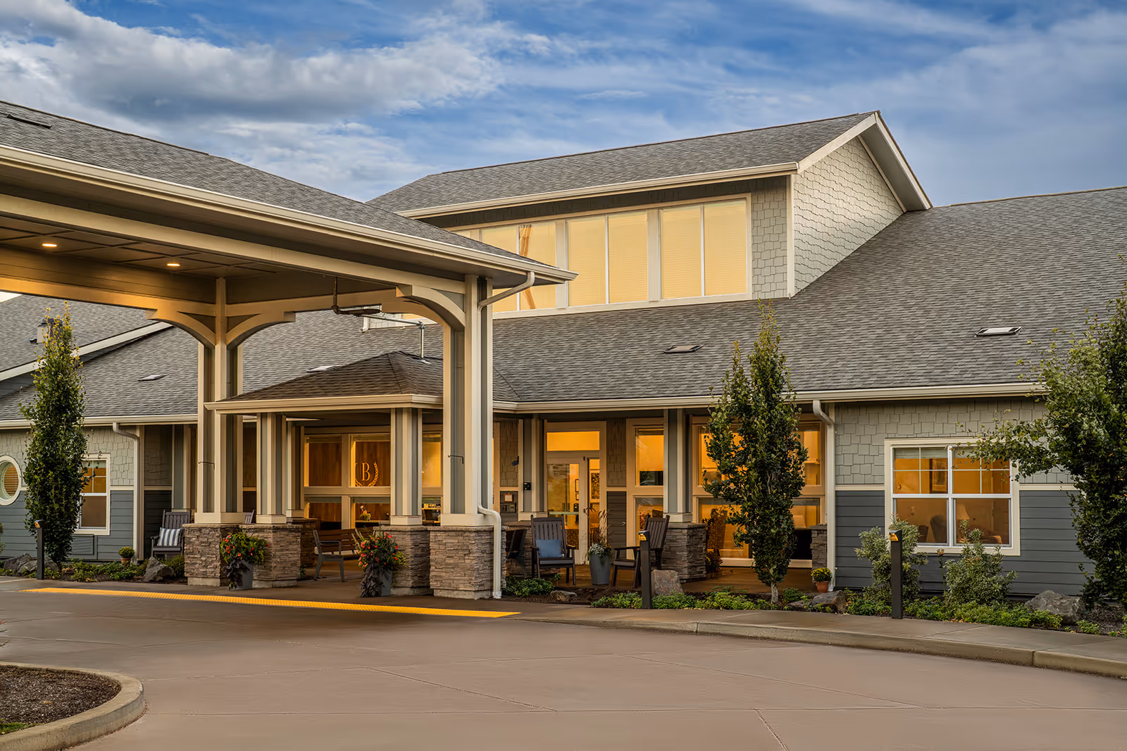 Exterior view of a senior living facility building with a covered entrance, stone pillars, and well-maintained landscaping under a partly cloudy sky during early evening.