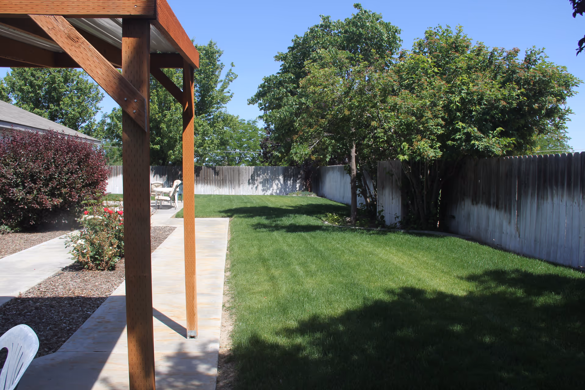 A sunny outdoor area at Ashley Manor Senior Living featuring a well-maintained green lawn, a wooden pergola structure, a concrete walkway, bushes with flowers, and a wooden fence surrounding the yard.