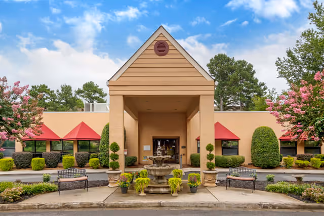 Front exterior view of a single-story building with a peaked entrance, beige walls, red awnings over windows, a central water fountain, benches, and landscaped greenery including bushes and flowering trees under a blue sky with some clouds.