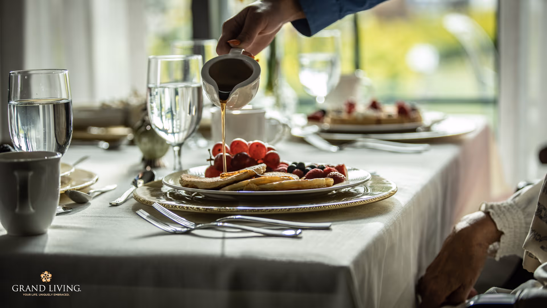 A close-up of a breakfast table with a plate of pancakes, grapes, and berries. A hand is pouring syrup onto the pancakes. The table is set with glasses of water, cups, and silverware, with a bright window in the background.