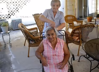 Two elderly women on a covered outdoor patio; one woman in a pink checkered dress is seated in a metal chair, while the other woman in a white patterned blouse stands behind her, touching her hair. The patio has several wicker chairs, a small table with a potted plant, and an air conditioning unit in the background.