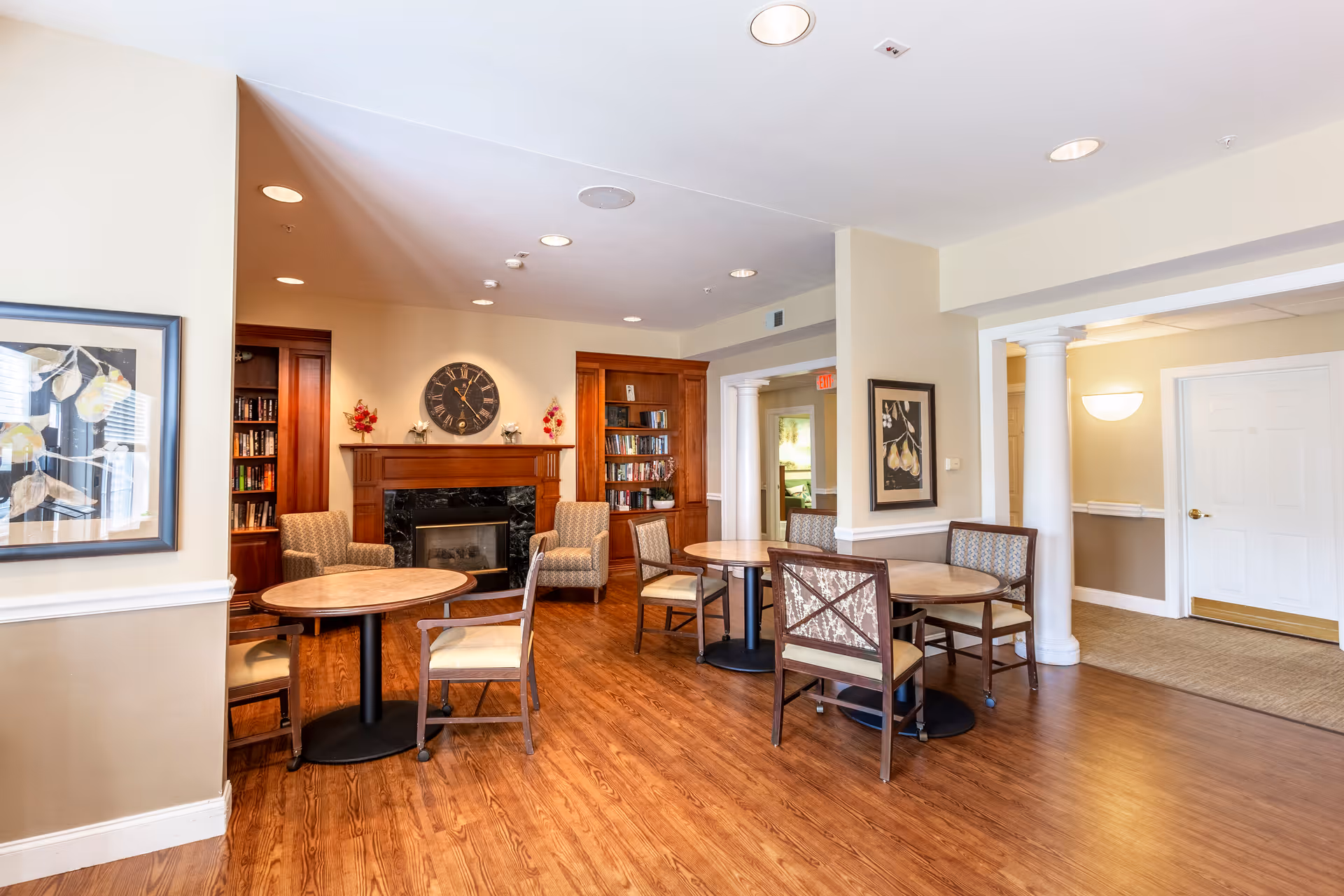 A cozy common area with wooden floors, round tables with chairs, a fireplace with a clock above it, built-in bookshelves, framed artwork on the walls, and soft lighting from ceiling fixtures and wall sconces.