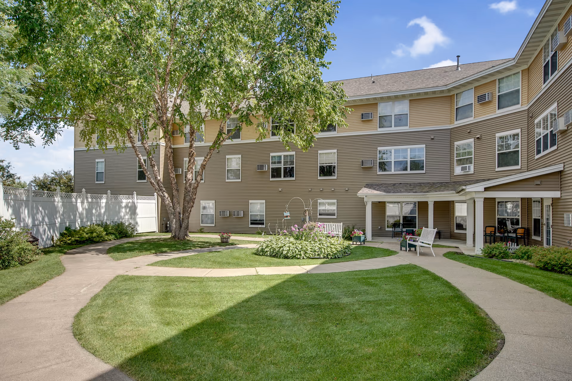 Sunlit courtyard with a circular walkway, lawn, benches, plants and a three-story assisted living building in the background.