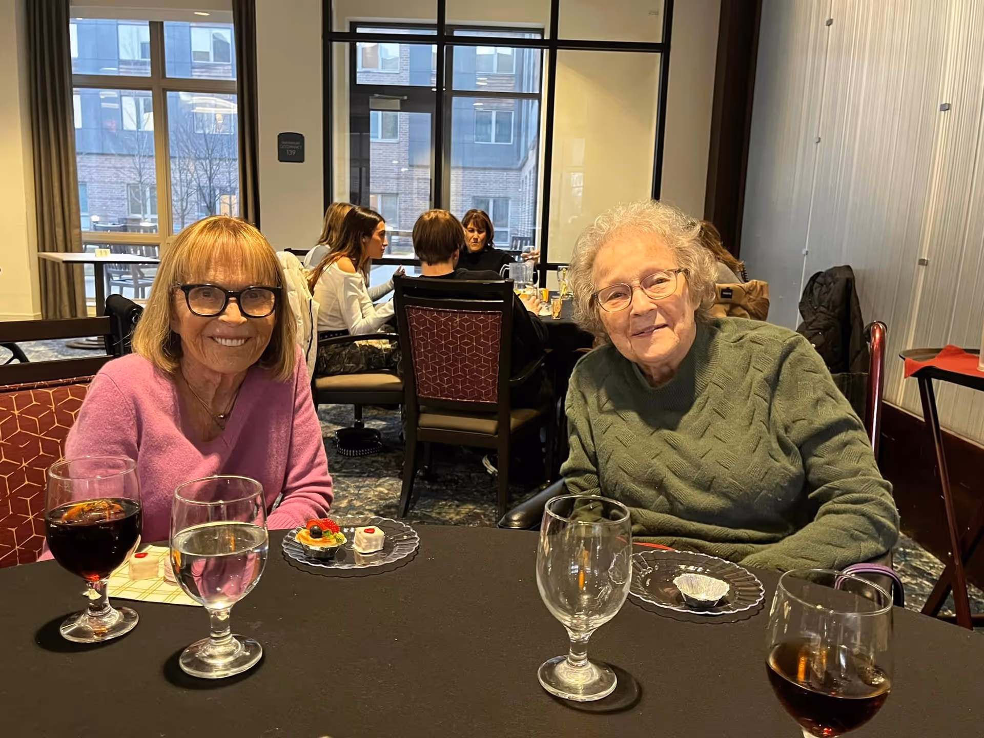 Two elderly women sit at a dining table with drinks and small desserts in a communal dining room.