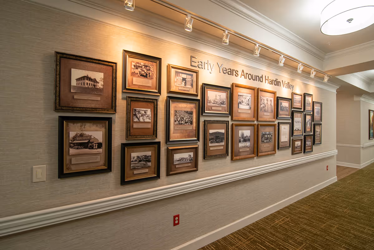 A hallway wall display featuring multiple framed historical photographs and documents under the title 'Early Years Around Hardin Valley'. The wall is well-lit with spotlights and the hallway has a green carpet and beige walls with white trim.