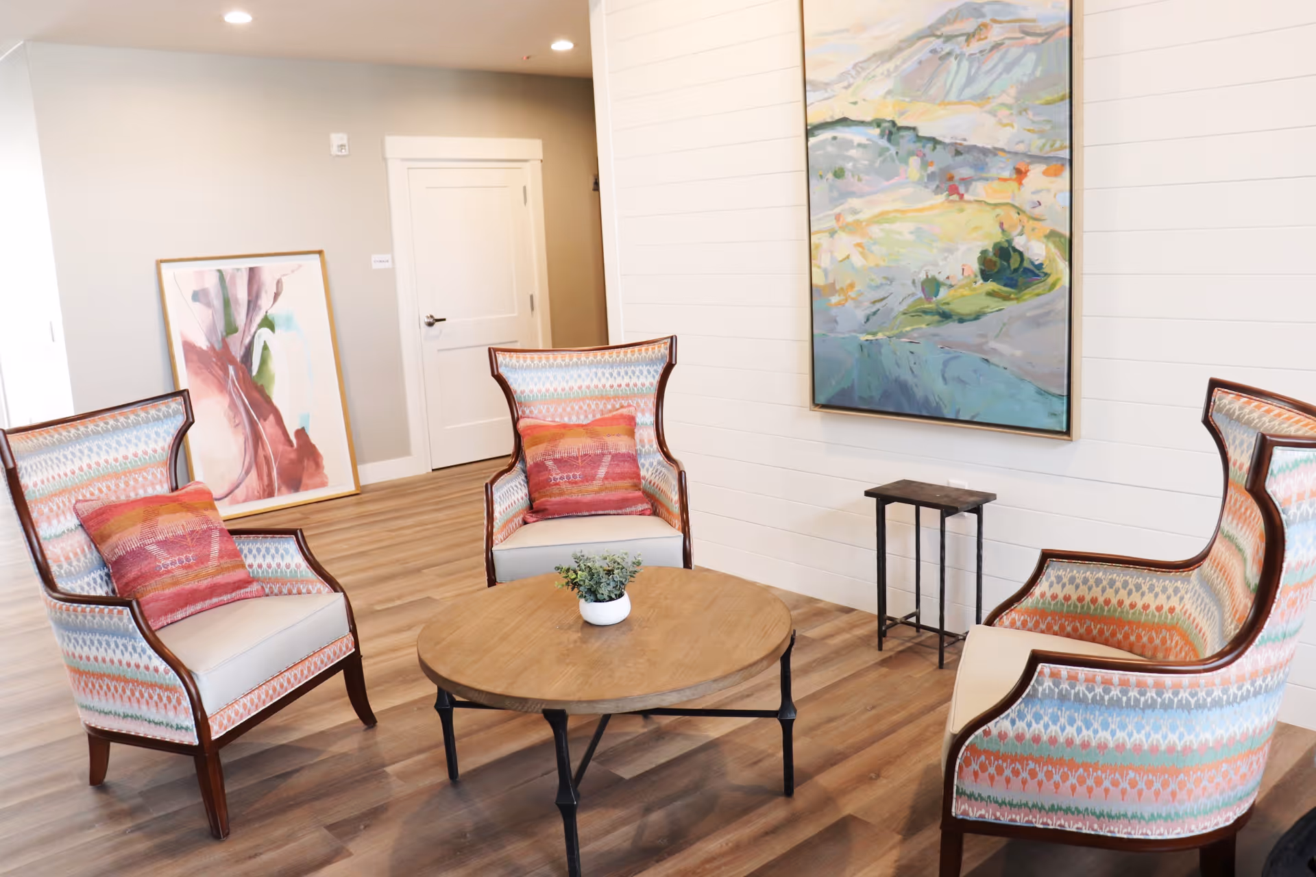 Bright lounge with three patterned armchairs arranged around a round wooden coffee table, decorative pillows, and wall artwork.