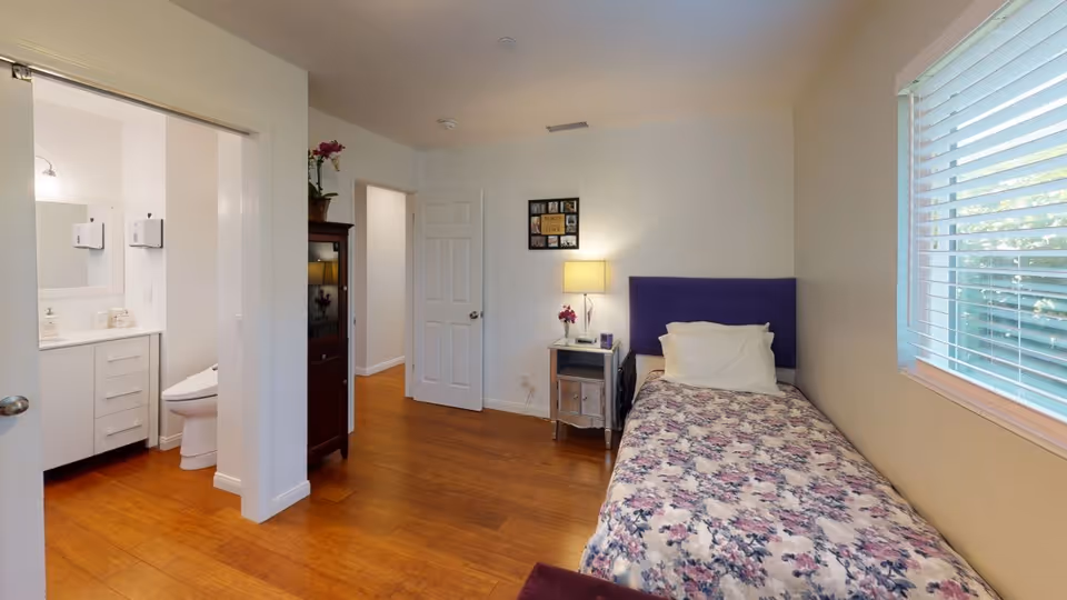 A small bedroom with a single bed covered in a floral bedspread, a purple headboard, and a white pillow. Next to the bed is a small nightstand with a lamp and a small vase with flowers. There is a window with white blinds on the right wall. The room has wooden flooring and white walls. To the left, there is an open doorway leading to a bathroom with a toilet and a sink visible. Another door is open, showing a hallway with wooden flooring.