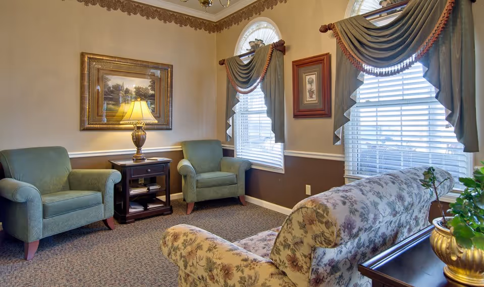 A cozy living room with two green armchairs, a floral patterned sofa, a wooden side table with a lamp, framed artwork on the walls, and two windows with decorative drapes and blinds.