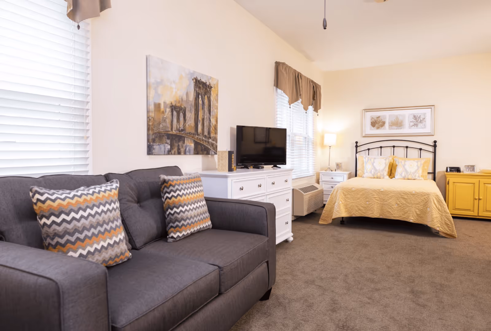 A bright studio room featuring a gray couch with patterned pillows, a TV on a white dresser, and a yellow-covered bed.