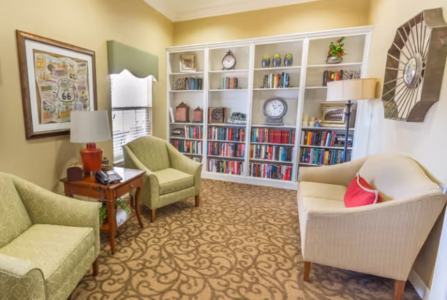 A cozy sitting area in an assisted living facility featuring two green armchairs and a beige loveseat with a red pillow arranged around a wooden side table with a lamp and telephone. Behind the seating is a large white bookshelf filled with books, decorative items, and clocks. The room has beige walls, a patterned carpet, a window with blinds and a green valance, and wall decorations including a framed Route 66 map and a large clock.