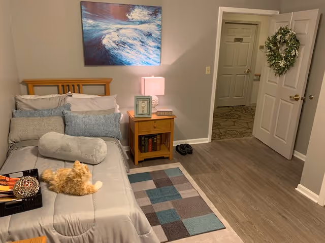 A cozy bedroom with a neatly made bed featuring multiple pillows and a stuffed animal cat on top. Next to the bed is a wooden nightstand with a lamp, picture frame, and books. The walls are painted light gray, and a painting of ocean waves hangs above the bed. The floor has a small patterned rug, and an open door reveals a hallway with carpeted flooring and another closed door. A decorative wreath hangs on the door to the right.