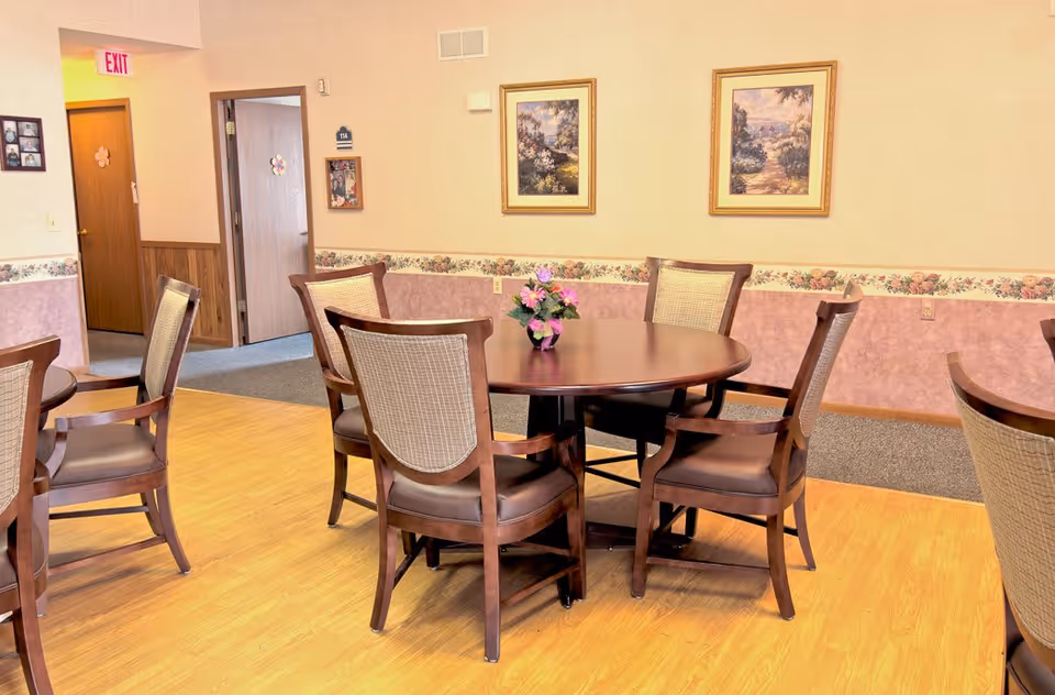 A dining area with a round wooden table surrounded by six wooden chairs with cushioned seats and backs. A small flower arrangement is placed in the center of the table. The room has light-colored walls with a floral wallpaper border and two framed landscape paintings. There is a doorway leading to another room and an exit sign above a door in the background.