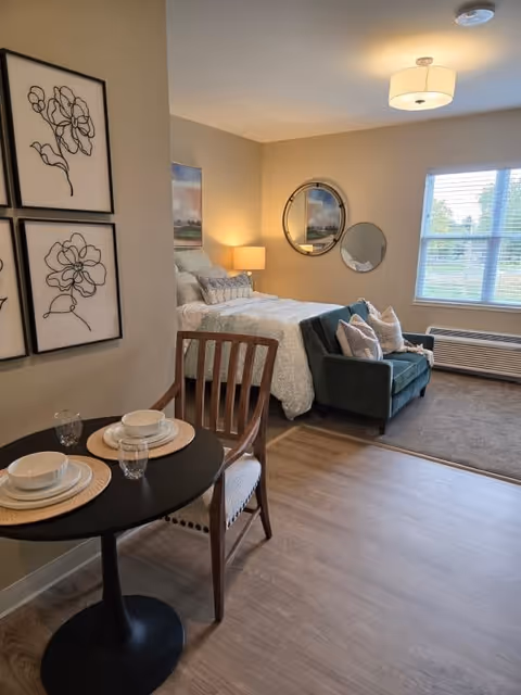 A cozy studio apartment interior featuring a bed with pillows and a side lamp, a small green loveseat with decorative pillows, a round black dining table set for two with plates and glasses, wooden chair, wall art with floral line drawings, two round mirrors on the wall, and a window with blinds letting in natural light.